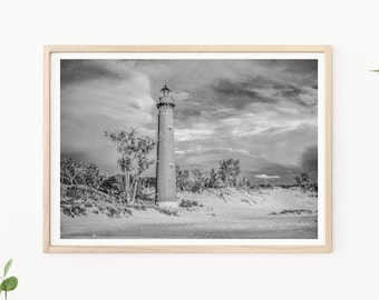 May include: A black and white photograph of a lighthouse on a sandy beach with a cloudy sky. The lighthouse is tall and slender, with a white top and a black base. The beach is empty, and the sky is filled with dark clouds.