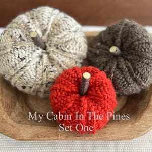 May include: Three knitted pumpkins in cream with black flecks, dark brown, and bright red, each topped with a small wooden stick stem. The pumpkins are arranged inside a shallow, rectangular wooden bowl. Text reads: My Cabin In The Pines Set One.