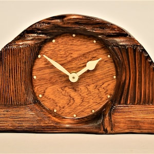 Puede incluir: Un reloj de madera con una esfera redonda y agujas blancas. El reloj está hecho de madera oscura y tiene una forma única y curvada.