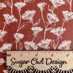 May include: Close-up of a fabric with a repeating pattern of white floral illustrations on a rust-colored background. A wooden ruler with the text "Sugar Owl Design" is placed over the fabric.