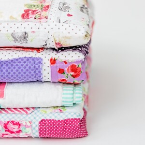 May include: A stack of five colourful patchwork quilts with floral and geometric patterns. The quilts are made of different fabrics and colours, including pink, purple, white and blue.