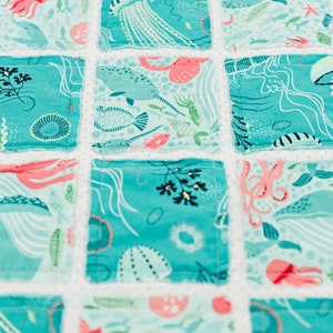 May include: A turquoise blue patchwork quilt with a white background. The quilt features a variety of sea creatures, including jellyfish, narwhals, and octopuses. The quilt is made of cotton fabric and is perfect for a nursery or child's room.