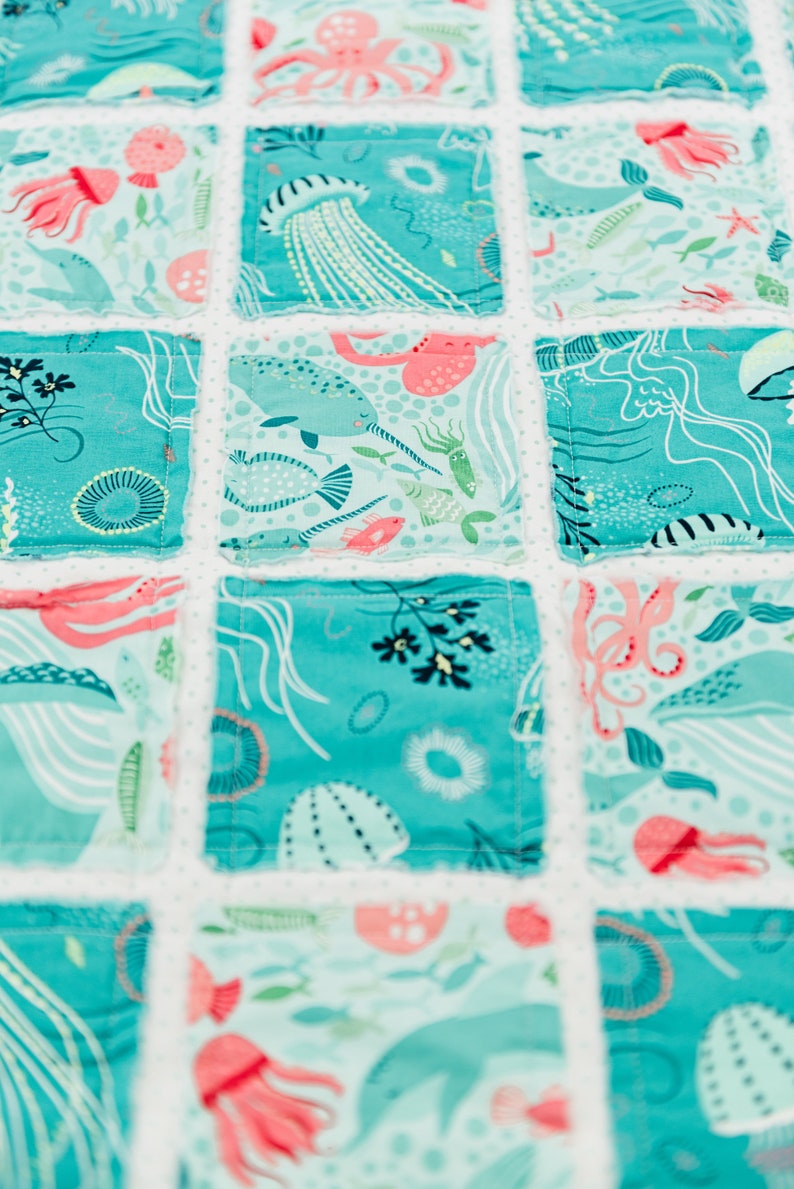 May include: A turquoise blue patchwork quilt with a white background. The quilt features a variety of sea creatures, including jellyfish, narwhals, and octopuses. The quilt is made of cotton fabric and is perfect for a nursery or child's room.
