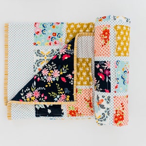 May include: A patchwork quilt featuring a range of floral patterns in shades of pink, blue, yellow, and red. The quilt is made up of squares and rectangles, with the edges trimmed in a yellow fabric. The quilt is rolled up and tied with a ribbon.