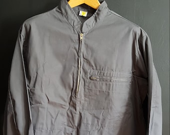 Può includere: Una camicia grigia a maniche lunghe con chiusura a zip e colletto alto. La camicia ha una tasca sul petto sinistro e un cordoncino alla fine delle maniche.