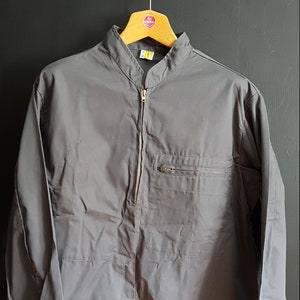 Peut inclure: Une chemise grise à manches longues avec une fermeture éclair et un col montant. La chemise a une poche sur la poitrine gauche et un cordon de serrage au bas des manches.