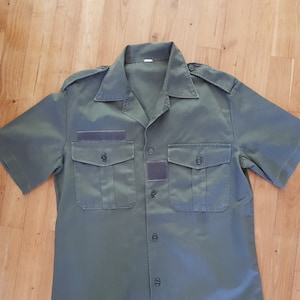 Può includere: Una camicia verde a maniche corte con bottoni, due tasche a patta e una toppa sulla tasca sinistra.