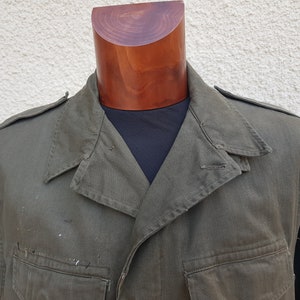 Pode incluir: Uma jaqueta militar verde com fecho de botões e gola pontiaguda. A jaqueta tem dois bolsos grandes na frente e um único bolso na manga esquerda.