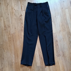 Può includere: Pantaloni neri con una striscia di raso sul lato. I pantaloni hanno la parte anteriore a pieghe e una gamba dritta. I pantaloni sono realizzati in un tessuto scuro e sono probabilmente per abbigliamento formale.