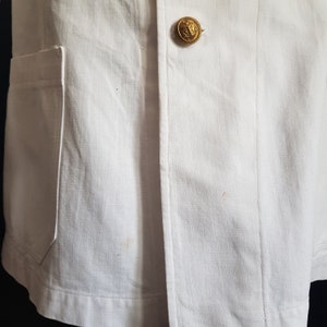 May include: Close-up of a white linen jacket with a gold button. The jacket has a pocket on the left side and a seam down the front.