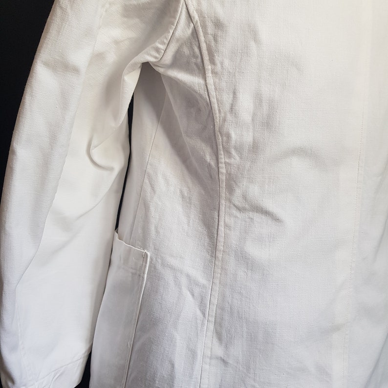 May include: White linen blazer with a single pocket on the left side. The blazer has a classic, tailored fit.