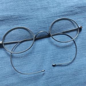 Könnte beinhalten: Silberfarbene Brille mit runden Gläsern und filigranem Drahtdesign. Die Brille hat einen Steg und Bügel, und die Gläser fehlen. Die Fassung befindet sich vor einem blauen Stoffhintergrund.