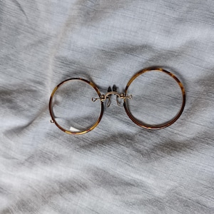 Può includere: Occhiali rotondi con montatura a fantasia tartarugata. Gli occhiali hanno un ponte e naselli color oro. Le lenti sono trasparenti. Gli occhiali sono appoggiati su un tessuto grigio chiaro.