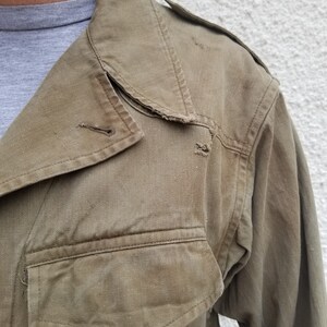 Może przedstawiać: Zbliżenie na brązową kurtkę z pł&oacute;tna z dużą kieszenią po lewej stronie. Kurtka ma zużyty, vintage'owy wygląd z widocznymi szwami i strzępionym kołnierzem.