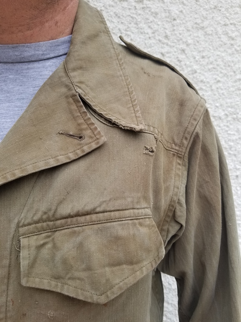 Może przedstawiać: Zbliżenie na brązową kurtkę z pł&oacute;tna z dużą kieszenią po lewej stronie. Kurtka ma zużyty, vintage'owy wygląd z widocznymi szwami i strzępionym kołnierzem.