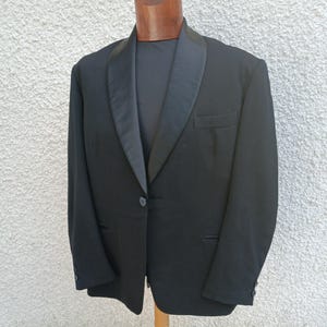Pode incluir: Casaco de smoking preto com lapela de cetim e fecho de um botão. O casaco tem um corte sob medida, um bolso no peito e punhos abotoados. A peça de roupa é exibida em um manequim de madeira.