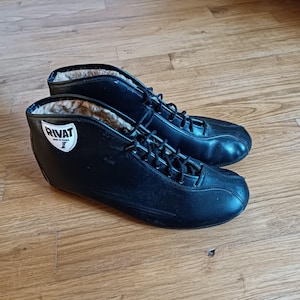 May include: A pair of black ice skates with laces. The skates have a high-top design with a white label that reads "RIVAT". The interior of the skates is lined with a light-colored material. The skates are on a wooden surface.