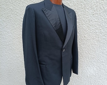 May include: A black tuxedo jacket featuring a single-button closure, satin-faced peak lapels, and a matching black shirt underneath. The garment is displayed on a mannequin torso, showcasing the structured shoulders and tailored fit of the piece.