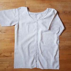 May include: A white linen shirt with a button-up front and short sleeves. The shirt is laid flat on a wooden surface.