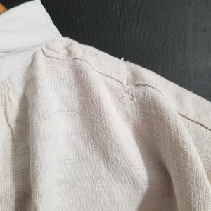 May include: A close-up of the shoulder seam of a white linen shirt. The fabric is textured and has a slight sheen.