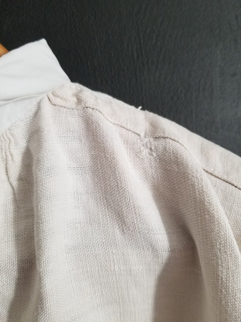 May include: A close-up of the shoulder seam of a white linen shirt. The fabric is textured and has a slight sheen.