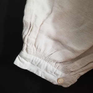 May include: Close-up of the cuff of a cream-colored, long-sleeved shirt with a single button closure.