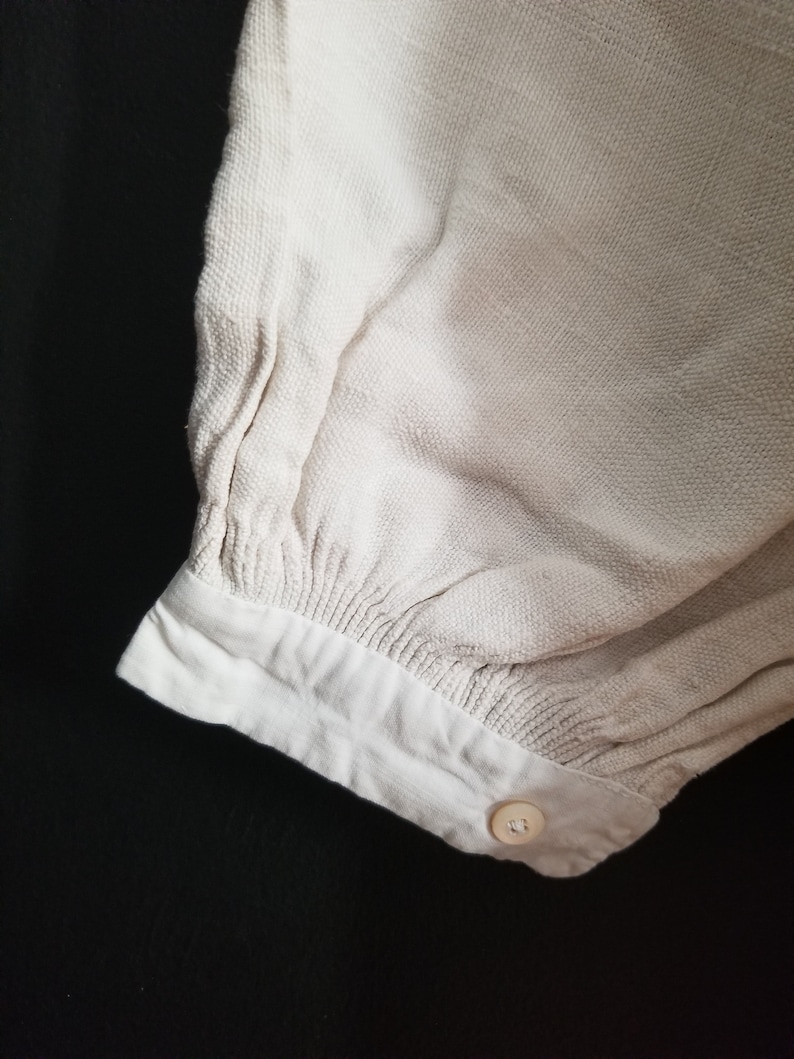 May include: Close-up of the cuff of a cream-colored, long-sleeved shirt with a single button closure.