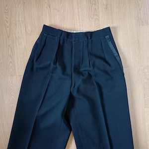Può includere: Pantaloni blu navy scuro con vita alta e parte anteriore a pieghe. I pantaloni hanno un design classico con gamba dritta e un dettaglio a righe in raso sul lato. Il tessuto sembra essere un materiale di peso medio, adatto per abbigliamento formale o semi-formale.