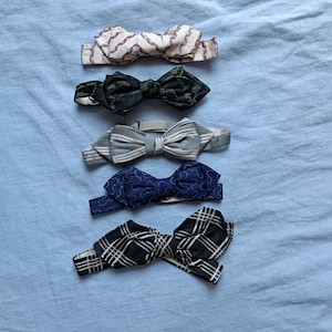 Può includere: Cinque papillon disposti verticalmente. Il papillon in alto è color crema con un motivo a zigzag rosso. Il secondo è verde scuro con una sottile stampa a foglie. Il terzo è grigio con strisce bianche. Il quarto è blu navy con pois bianchi. L'ultimo è a quadri bianchi e neri.