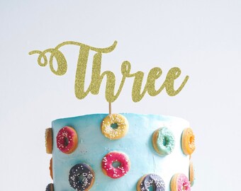 May include: A blue frosted cake decorated with colorful donuts and a gold glitter topper that says "Three".