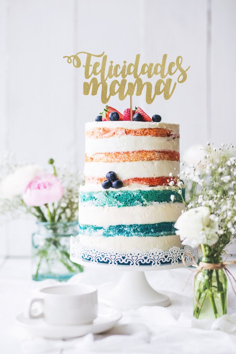 May include: A two-tiered cake decorated with white frosting and colorful layers. The cake is topped with fresh berries and a gold glitter cake topper that reads "felicidades mama".