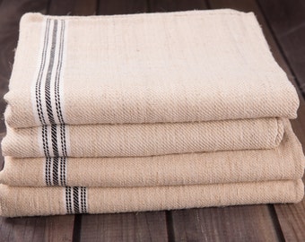 May include: A stack of four beige kitchen towels with a woven texture. Each towel has a black and white striped border along one edge. The towels are neatly folded and stacked on a dark wooden surface.