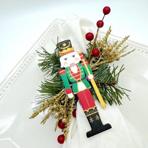 May include: A wooden nutcracker napkin ring with a red, green, and black design. The nutcracker is holding a gold stick and is surrounded by green pine branches, red berries, and gold glitter.