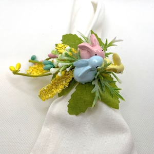 May include: A white napkin ring adorned with a spring-themed arrangement. The arrangement features three pastel-colored bunny figurines, green leaves, and yellow floral accents. A charming Easter decoration.