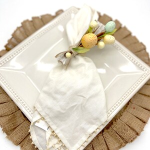 May include: A white napkin with a beaded edge is tied with a sprig of faux Easter eggs in shades of yellow, green, and blue. The napkin is placed on a white square plate with a beaded border, which is set on a brown burlap placemat.