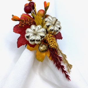 May include: A silver pumpkin napkin ring with fall foliage and berries. The ring is decorated with red, orange, and yellow leaves, as well as small red berries and yellow berries.