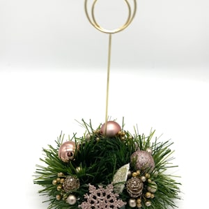 May include: A gold metal table number holder with a double ring design sits on a small green wreath decorated with pink and gold ornaments and a pink snowflake.