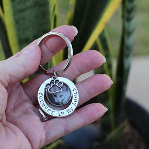 May include: A silver-colored keychain with a circular pendant featuring a cat photo. The pendant has a paw print design and the words "forever in my heart" around the cat's image. The keychain is held in a hand.