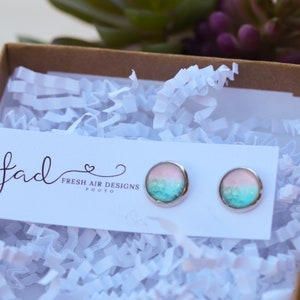 May include: A pair of silver stud earrings with a pink and blue ombre design. The earrings are displayed on a white card with the text "Fresh Air Designs Photo".