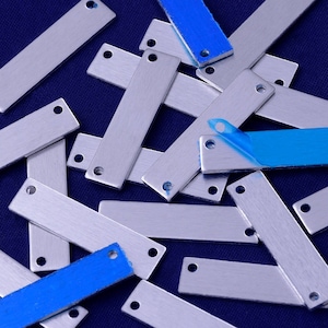 Könnte beinhalten: Ein Haufen rechteckiger Anhänger aus silbernem Metall mit jeweils zwei Löchern. Die Anhänger liegen auf einer dunkelblauen Oberfläche verstreut. Einige der Anhänger sind mit blauem Klebeband beklebt.