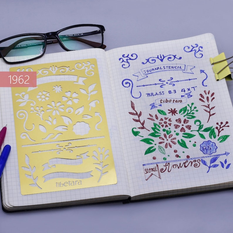 May include: A gold metal stencil with floral and banner designs. The stencil is labeled "tibetara" and is 0.3 x 4 inches. The stencil is on a page of a notebook with the words "JOURNAL STENCIL" and "stencil flowers" written in blue ink.