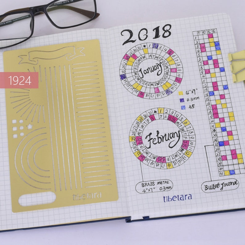 May include: A brass metal stencil with various shapes and lines, including a banner, sun rays, circles, and a grid. The stencil is 4 inches by 7 inches and 0.3 millimeters thick. The stencil is labeled "Tibetara" and is used for bullet journaling.