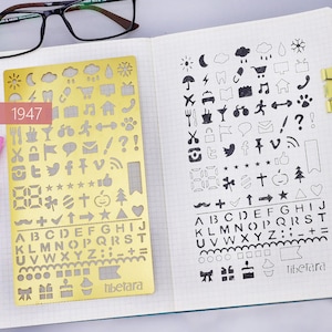 May include: A gold metal stencil with various symbols, letters, and numbers. The stencil is on a white gridded notebook page with the same symbols, letters, and numbers drawn in black ink. The stencil has the number 1947 printed on it and the word "tibetara" at the bottom.