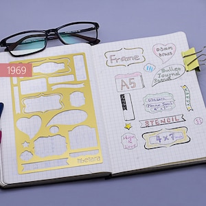 May include: A gold stencil with various shapes, including rectangles, circles, hearts, and banners. The stencil is on a notebook page with handwritten notes, including "Frame", "A5", "Bullet Journal Spread", "Tibetara Planner Stencil 4 x 7", "Stencil 4 x 7 inch", and "Happy".