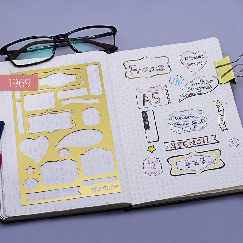 May include: A gold stencil with various shapes, including rectangles, circles, hearts, and banners. The stencil is on a notebook page with handwritten notes, including "Frame", "A5", "Bullet Journal Spread", "Tibetara Planner Stencil 4 x 7", "Stencil 4 x 7 inch", and "Happy".