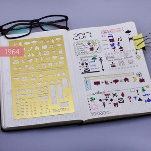 May include: A gold metal stencil with various shapes and symbols, including arrows, stars, hearts, and geometric shapes. The stencil is labeled "Tibetara" and is open on a page of a gridded notebook. The notebook page has handwritten notes and drawings in various colors.