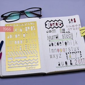May include: A gold metal stencil with a variety of shapes and letters, including numbers, alphabet letters, and symbols. The stencil is on a white gridded notebook page with handwritten text that says "tibetara", "03 mm", "journal stecil", "abcdefghijk", "Imnopqrstuv", "Wxyz", "47", "aug & 2017", and "$9.8".
