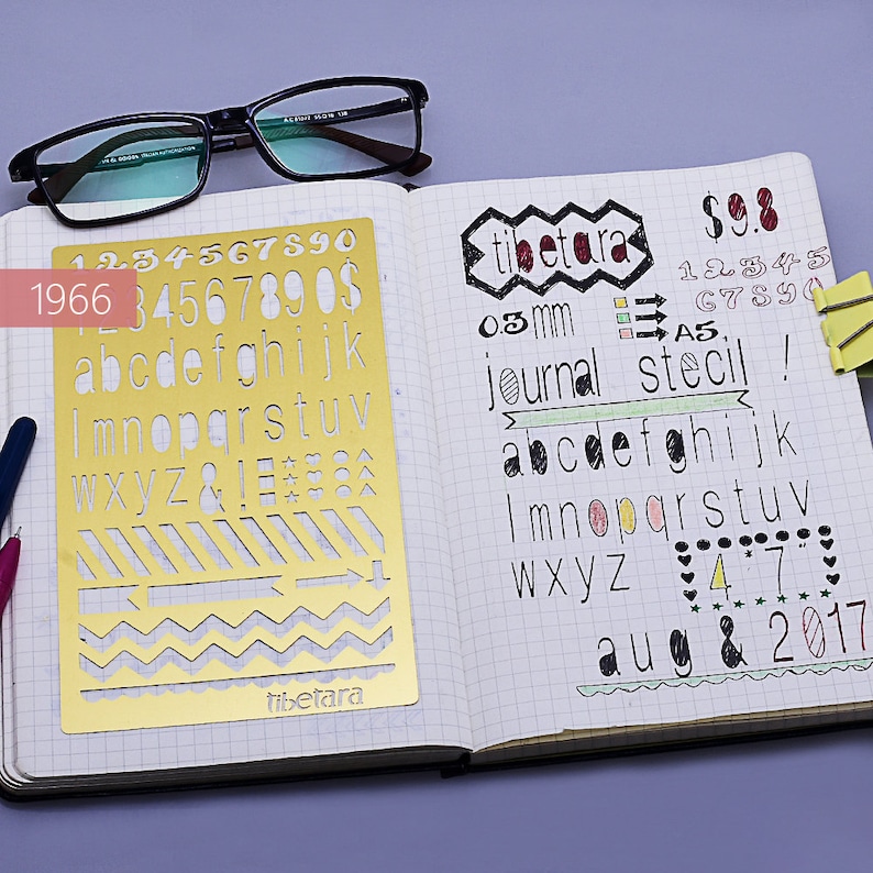 May include: A gold metal stencil with a variety of shapes and letters, including numbers, alphabet letters, and symbols. The stencil is on a white gridded notebook page with handwritten text that says "tibetara", "03 mm", "journal stecil", "abcdefghijk", "Imnopqrstuv", "Wxyz", "47", "aug & 2017", and "$9.8".