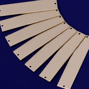 May include: A set of ten gold-colored metal rectangular tags with rounded corners and two holes at the top. The tags are arranged in a fan shape on a blue background.