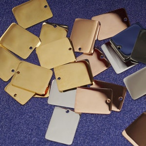 May include: Assortment of metal blank tags in various colors, including gold, silver, and rose gold. The tags are rectangular and have a hole at the top for attaching a string or ribbon.